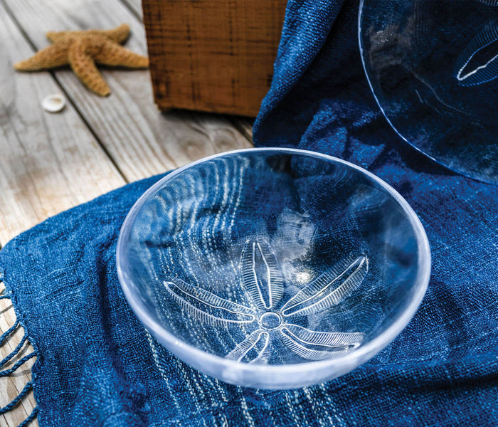 Clear glass bowl with white sand dollar design etched inside, resting on blue fabric.