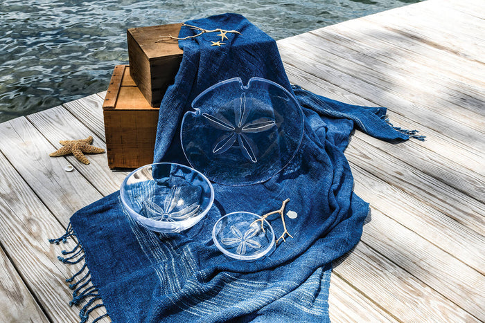 White ceramic plate with sand dollar relief design on blue fabric beside wooden boxes on a dock.