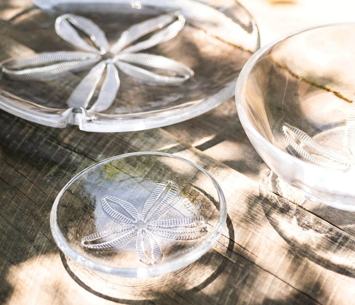 Clear glass platters with embossed sand dollar design, arranged on wooden surface in sunlight.