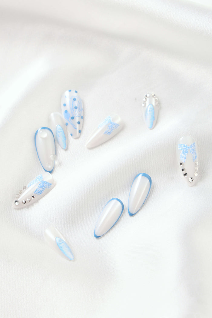 Almond press-on nails with blue polka dots, blue bows, and rhinestones on white satin.