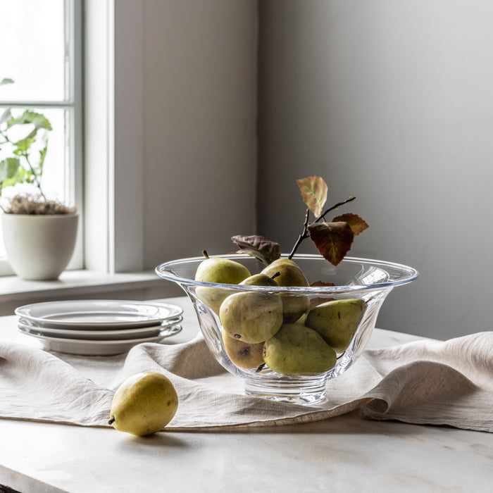 Clear glass bowl with flared rim filled with pears and decorative foliage on white linen table.