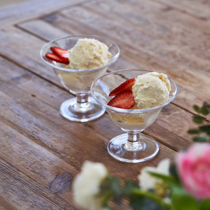Two clear glass dessert bowls with pedestal bases filled with vanilla ice cream and strawberry slices on wooden surface.