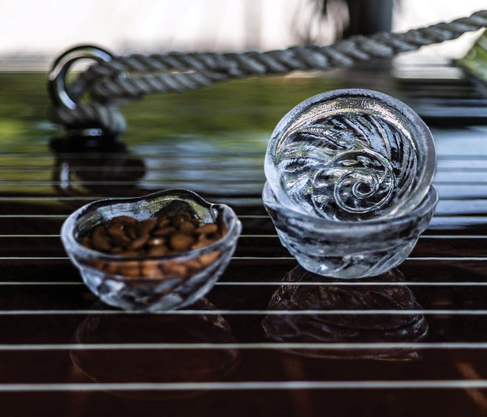 Small shell bowl with embossed wave design, displayed on dark striped surface with rope detail.