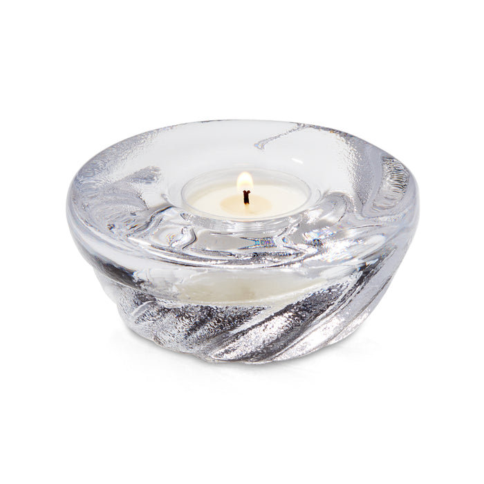Clear glass shell-shaped tealight holder with lit cream candle inside.