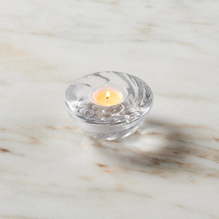 Clear glass shell-shaped tealight holder with lit cream-colored candle on marble surface.