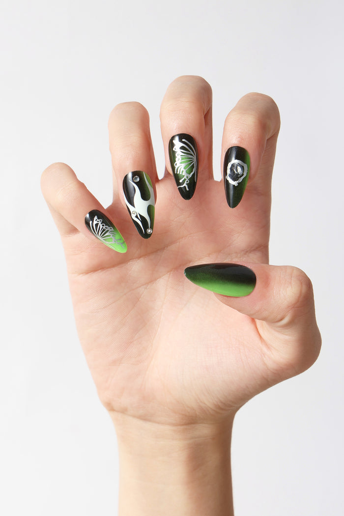 Woman's hand with SHE'S A BRAT press-on nails: black & green gradient, butterfly & flame designs, rhinestones.