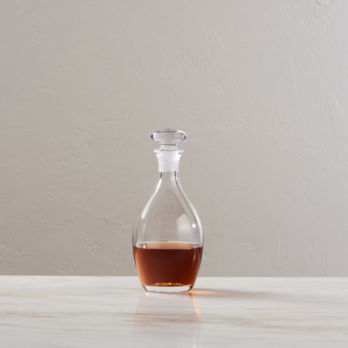 Clear glass decanter with stopper containing amber liquid, on white surface against neutral background.
