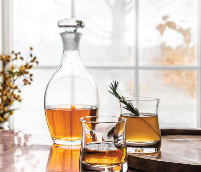 Clear glass decanter with stopper filled with amber liquid, accompanied by two drinking glasses with rosemary sprigs.