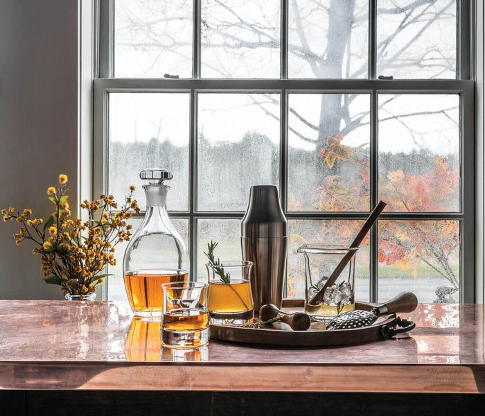 Clear glass decanter with stopper filled with amber liquid, displayed on table with matching glasses and bar tools.
