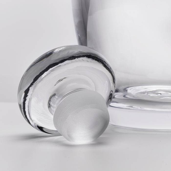 Clear glass decanter with spherical stopper and curved pouring spout, shown tilted on white surface.