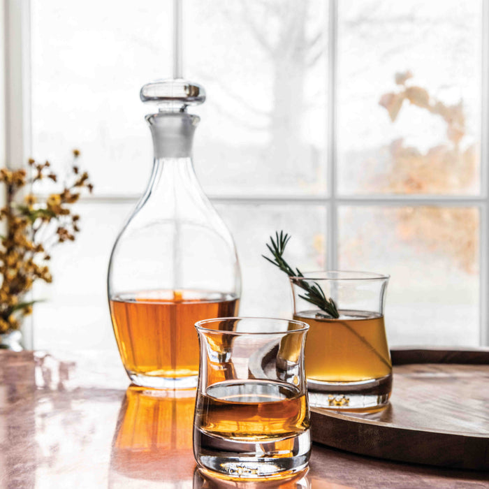 Two clear double old-fashioned glasses with amber liquid and rosemary sprigs next to a matching decanter with stopper.