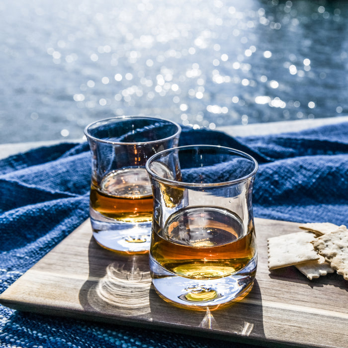 Two whiskey glasses with amber liquid on a wooden tray beside blue fabric and coastal background.