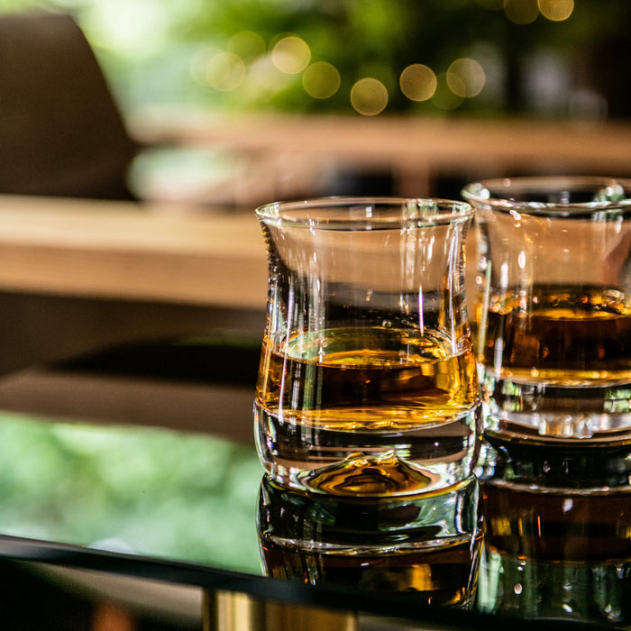 Two whiskey glasses filled with amber liquid on a dark surface with blurred outdoor background.