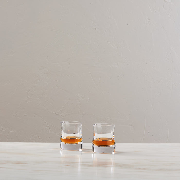 Two clear whiskey glasses filled with amber liquid on a white marble surface against a textured beige wall.