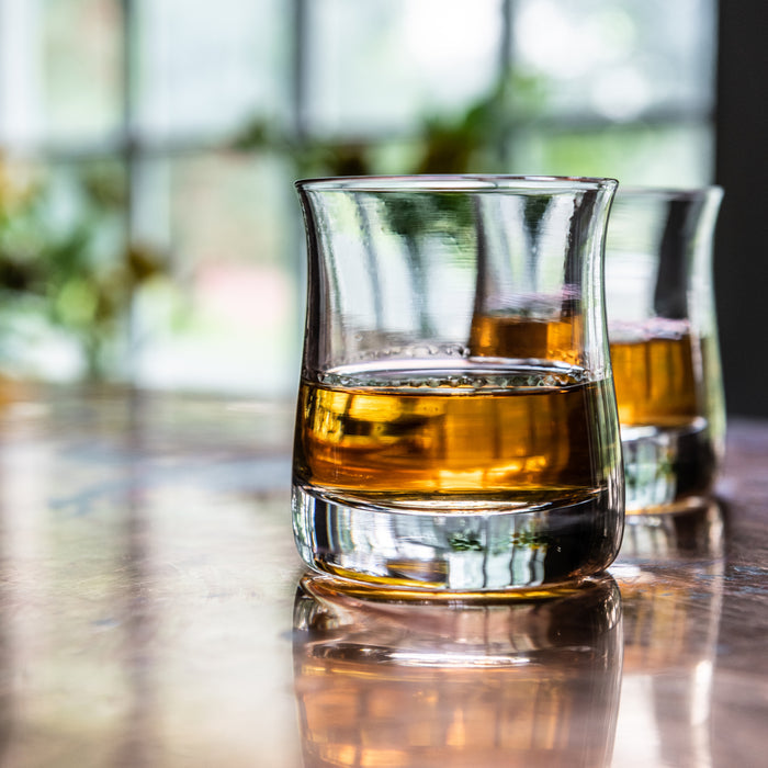 Two clear glasses containing amber whiskey, displayed on a reflective surface with a blurred window background.