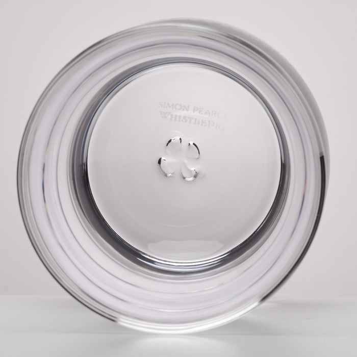 A clear circular glass photographed on a light gray background. Faintly etched text near the top reads 'SIMON PEARCE WHISTLE PIG'.