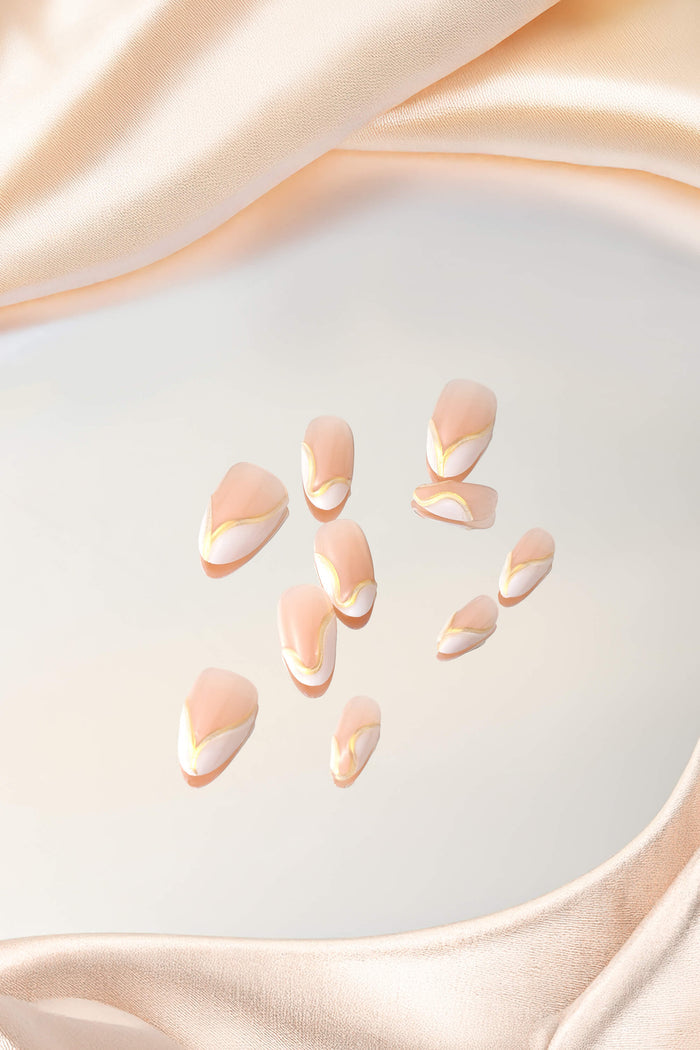 Short almond press-on nails with a nude base, white tips, and thin gold accents, set against a backdrop of draped satin.