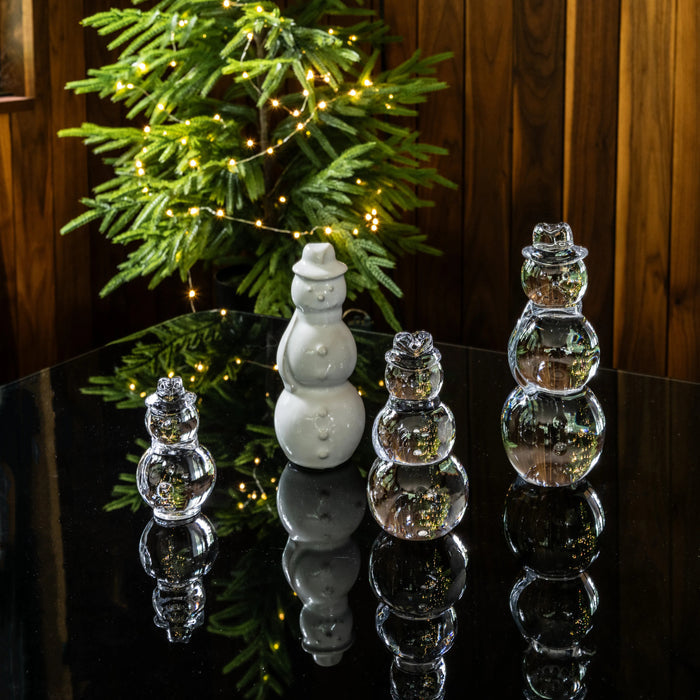 White ceramic snowman and three clear glass snowman figurines arranged on black surface with lit evergreen garland background