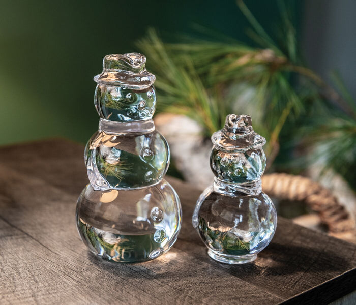 Clear glass snowman figurines with stacked spheres and hat, shown in two sizes next to evergreen branches.
