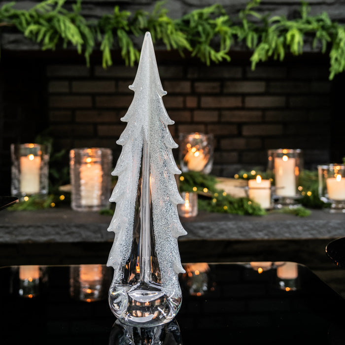 White frosted tree-shaped glass decor with textured finish and clear glass base, surrounded by candles and greenery.