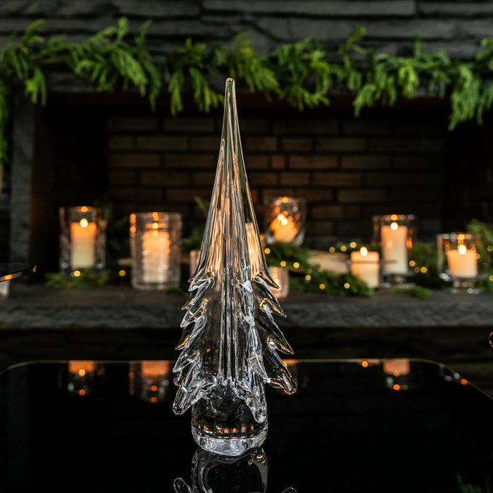 Clear glass tree-shaped decorative object with pointed top and tapered branches, displayed with candles and greenery.