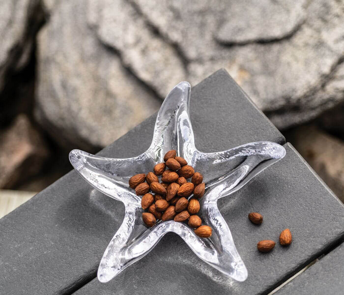 Starfish-shaped dish with clear glass and white accents holding roasted almonds on gray surface.