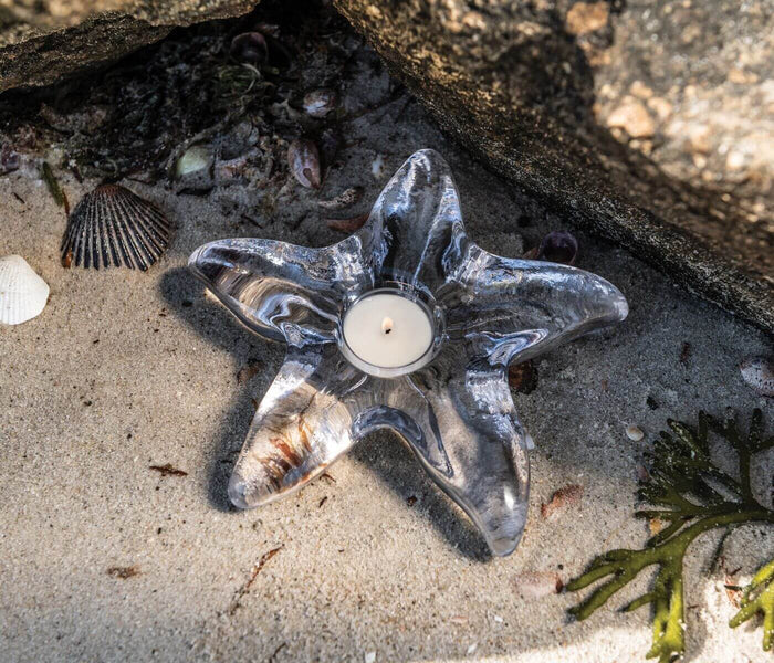Starfish-shaped tealight holder in dark iridescent glass with white candle, surrounded by shells and sand.