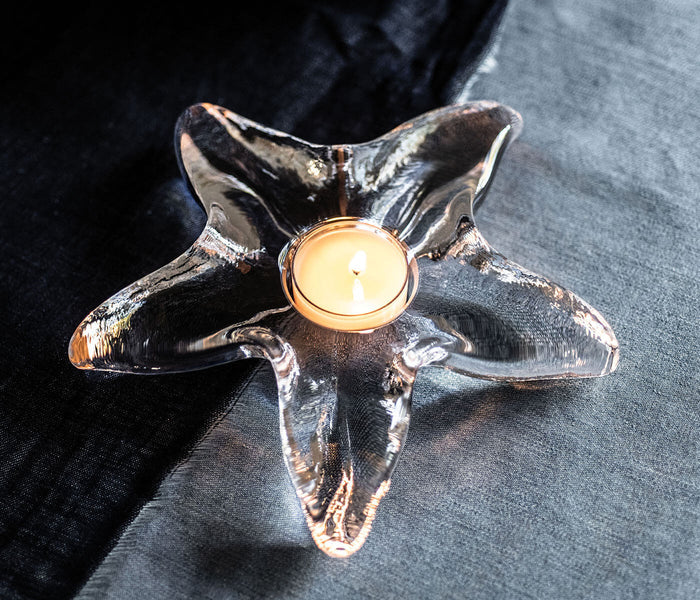 Clear glass starfish-shaped tealight holder with lit cream candle in center, displayed on dark and light fabric.