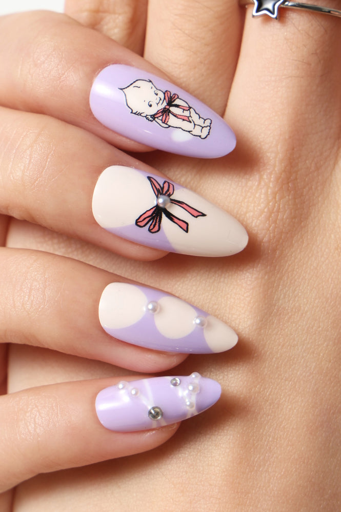 Purple press-on nails with Kewpie character design, bows, and pearl embellishments.