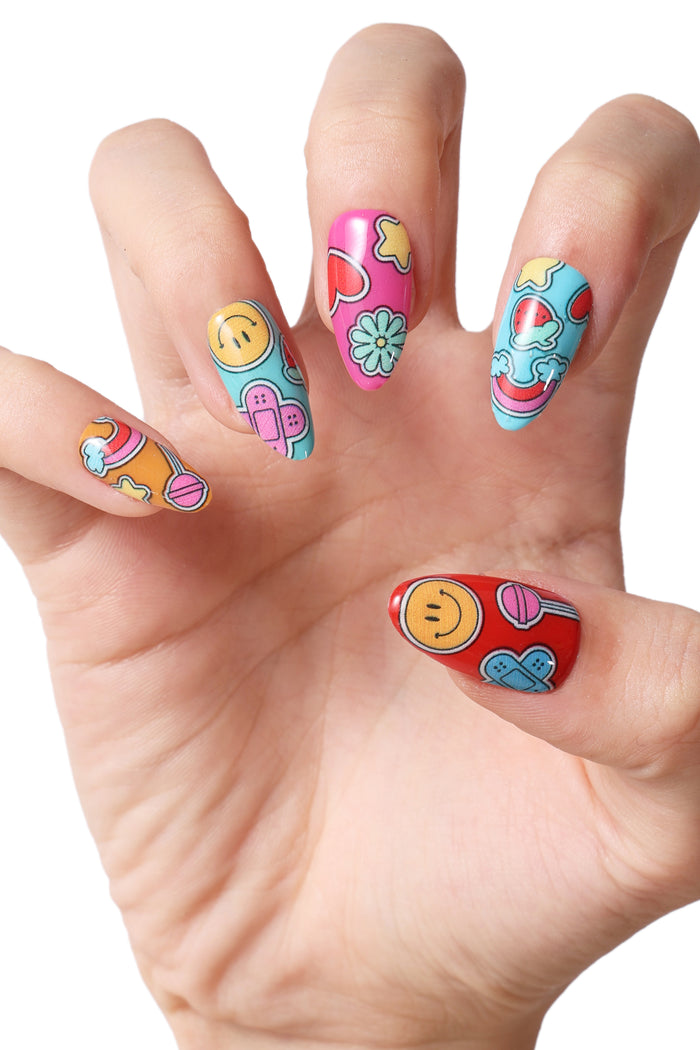 Press-on nails by Mercedes Bazan featuring colorful cartoon designs: smiley faces, rainbows, flowers, and band-aids.