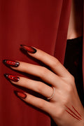 Almond press-on nails with a glossy crimson ombre and crystal chain accents, on a hand wearing a pearl ring.