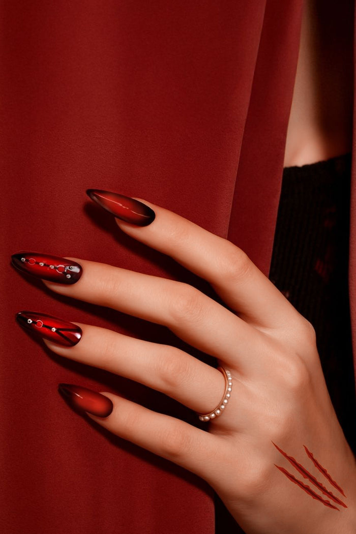 Almond press-on nails with a glossy crimson ombre and crystal chain accents, on a hand wearing a pearl ring.