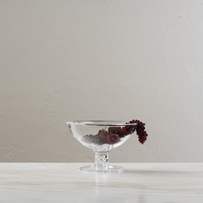 A clear glass pedestal bowl with a stem base, shown on a light marble surface against a textured beige wall. The bowl contains dark red berries, with a few resting on the rim.