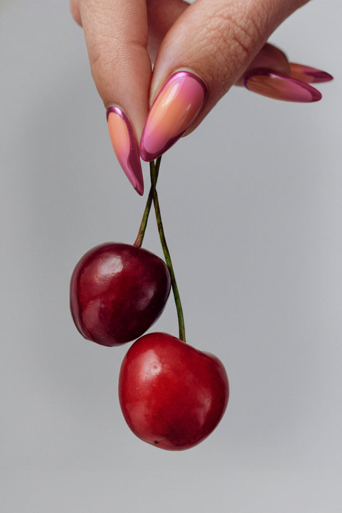 Hand with gradient sunset chrome press-on nails holding two ripe cherries.