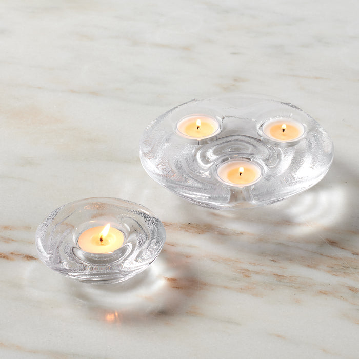 Clear glass tealight holders with lit cream-colored candles, one holding single candle and one holding three candles.