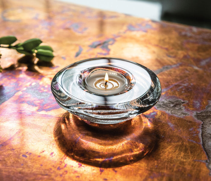 Glass tealight holder with lit candle on patterned surface next to green foliage.