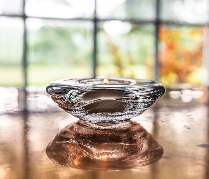 Clear glass tealight holder with organic wavy shape on a wooden surface near a window.