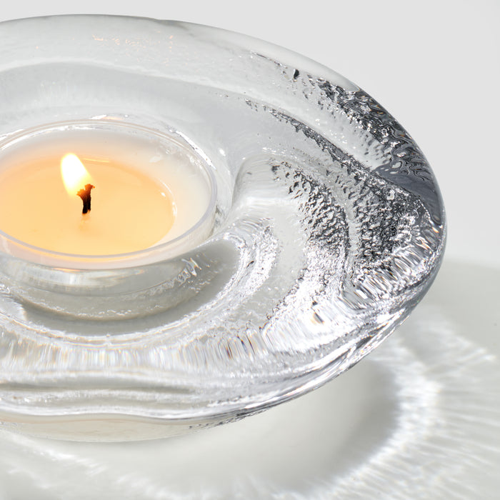 Lit cream tealight candle in clear glass holder with textured decorative rim.