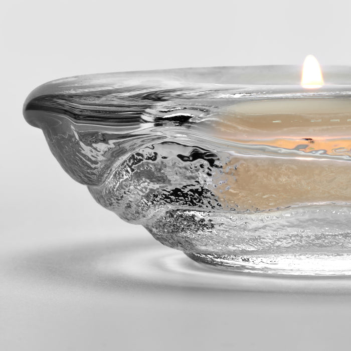 Clear glass tealight holder with molten textured rim containing lit cream-colored candle with flame.