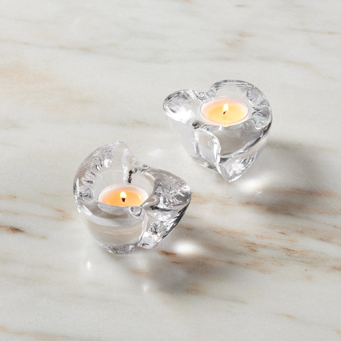 Two clear glass heart-shaped tealight holders with lit cream candles on marble surface.