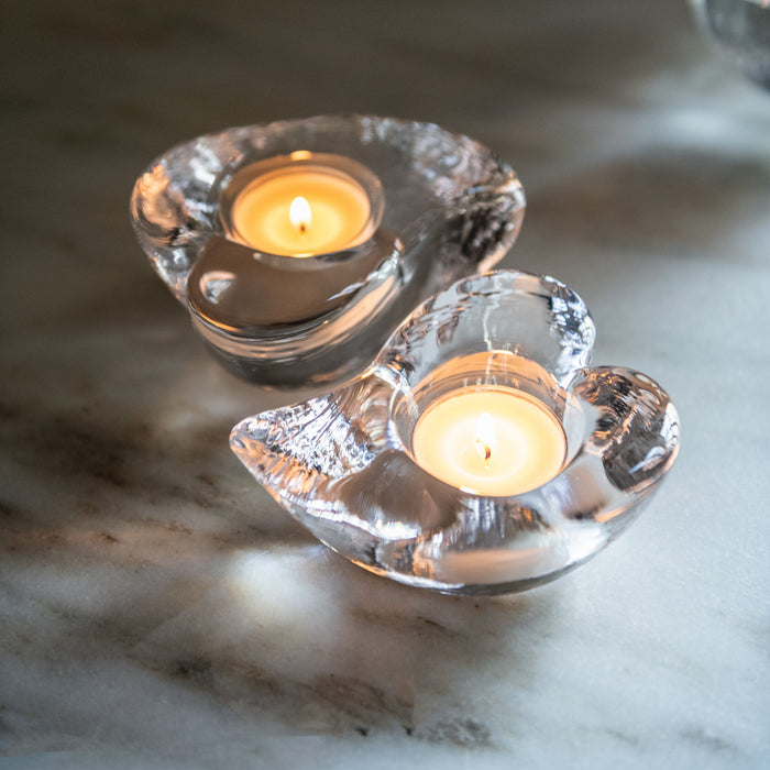 Two clear glass heart-shaped tealight holders with lit cream-colored candles on marble surface.