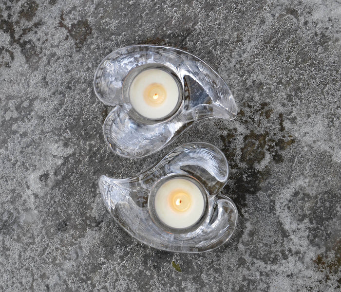 Two clear heart-shaped tealight holders with white candles and yellow flames inside, arranged vertically on concrete surface.