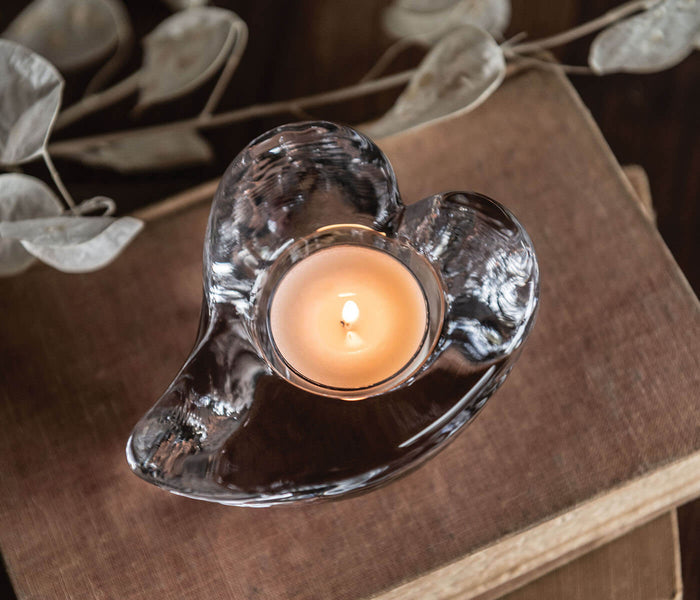 Heart-shaped clear glass tealight holder with lit cream-colored candle on tan fabric surface.
