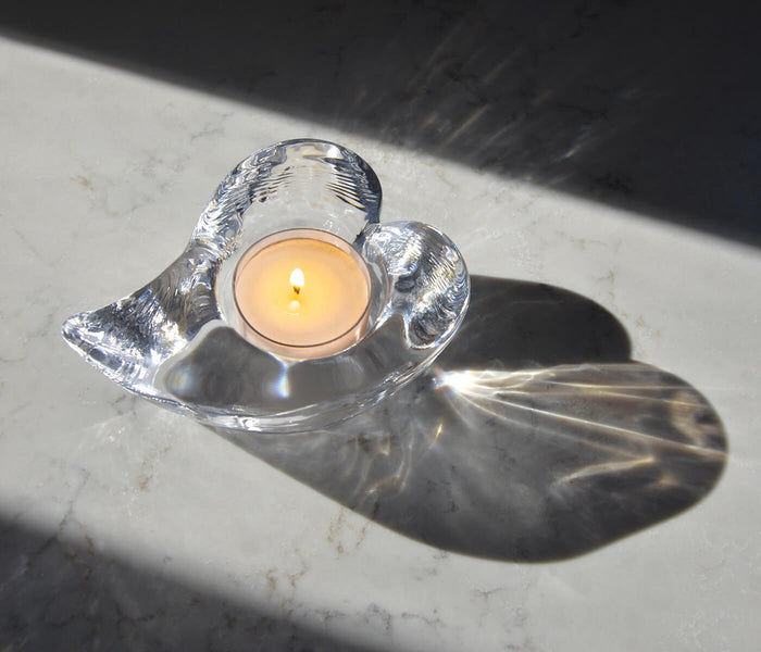 Clear glass heart-shaped tealight holder with lit cream candle inside, casting shadow on marble surface.