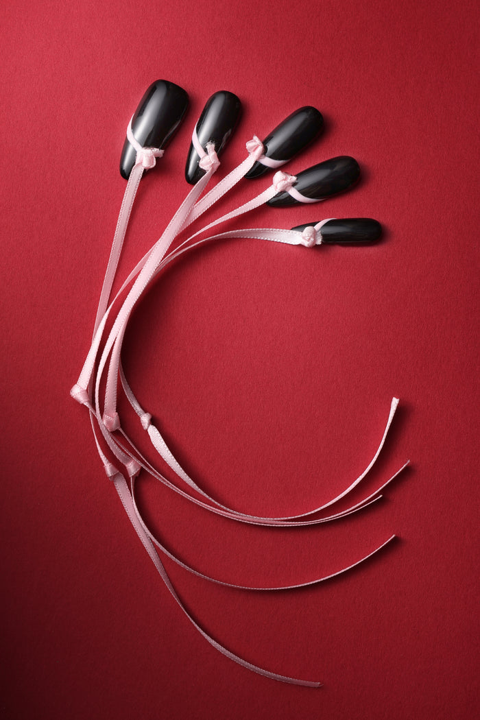 Black press-on nails with a delicate pink ribbon tied around each, arranged on a red background.