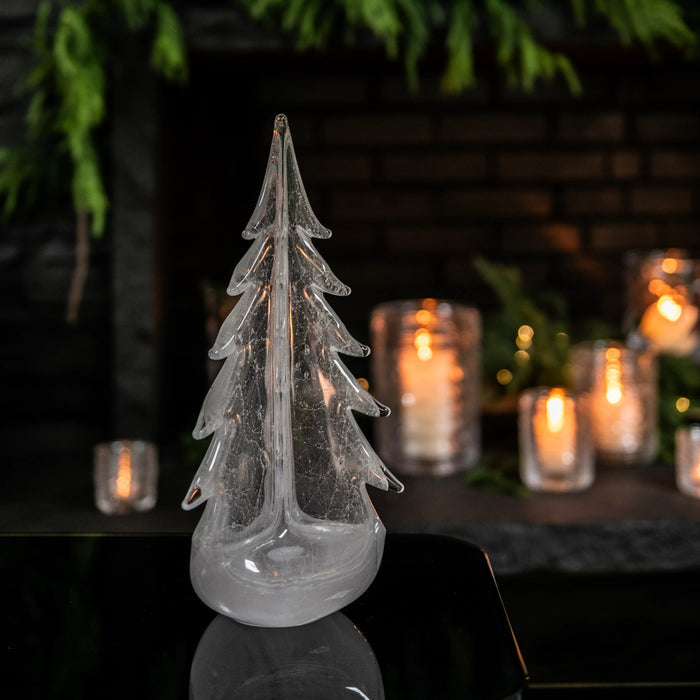 Clear glass evergreen tree candle with tiered branches and rounded base, displayed with lit candles and greenery in backgroun