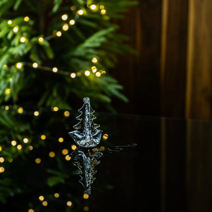 Clear glass tree-shaped ornament with gold accents, displayed in front of evergreen branches and warm white lights.