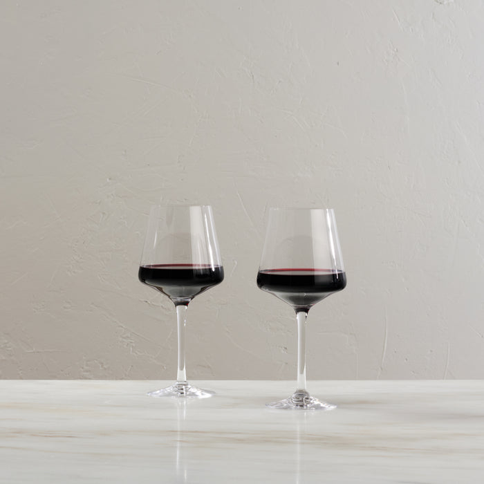 Two wine glasses filled with red wine on a white surface against a textured gray wall.