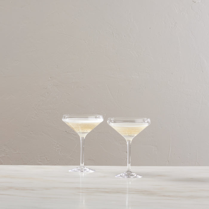 Two clear coupe glasses filled with pale yellow wine on a white surface against a neutral background.