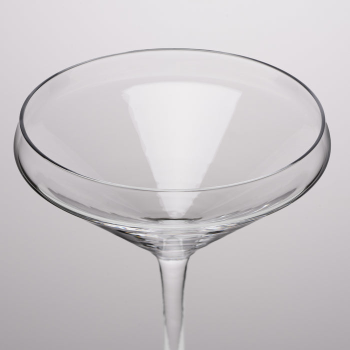 A clear glass coupe glass with a wide, shallow bowl and a slender stem, shown against a neutral light gray background.